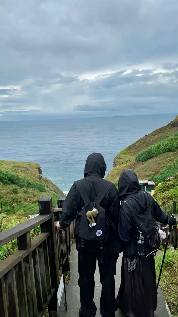 Couple hiking with backpacks and trekking poles down a wooden cliffside staircase overlooking the ocean.