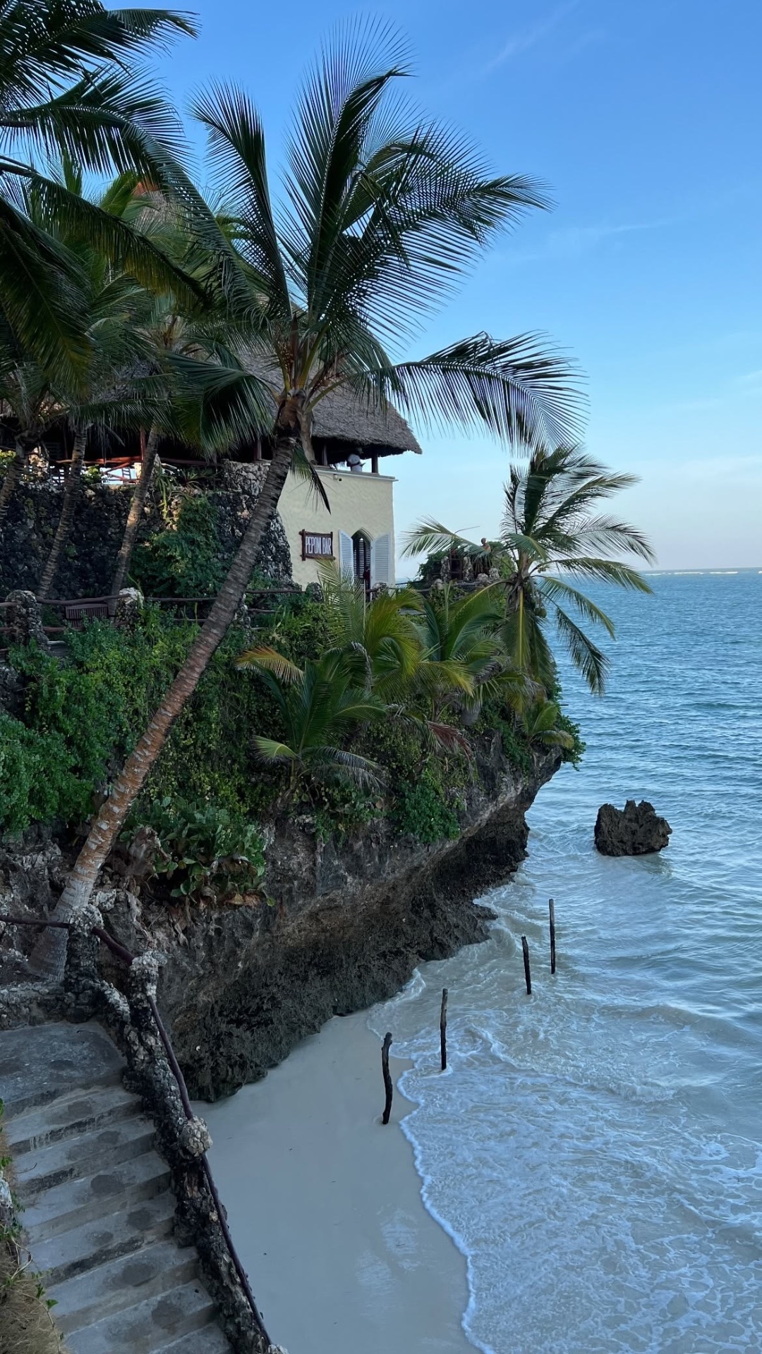 Palm-covered coral cliff with a cliffside bar above a white-sand beach and turquoise Indian Ocean surf.