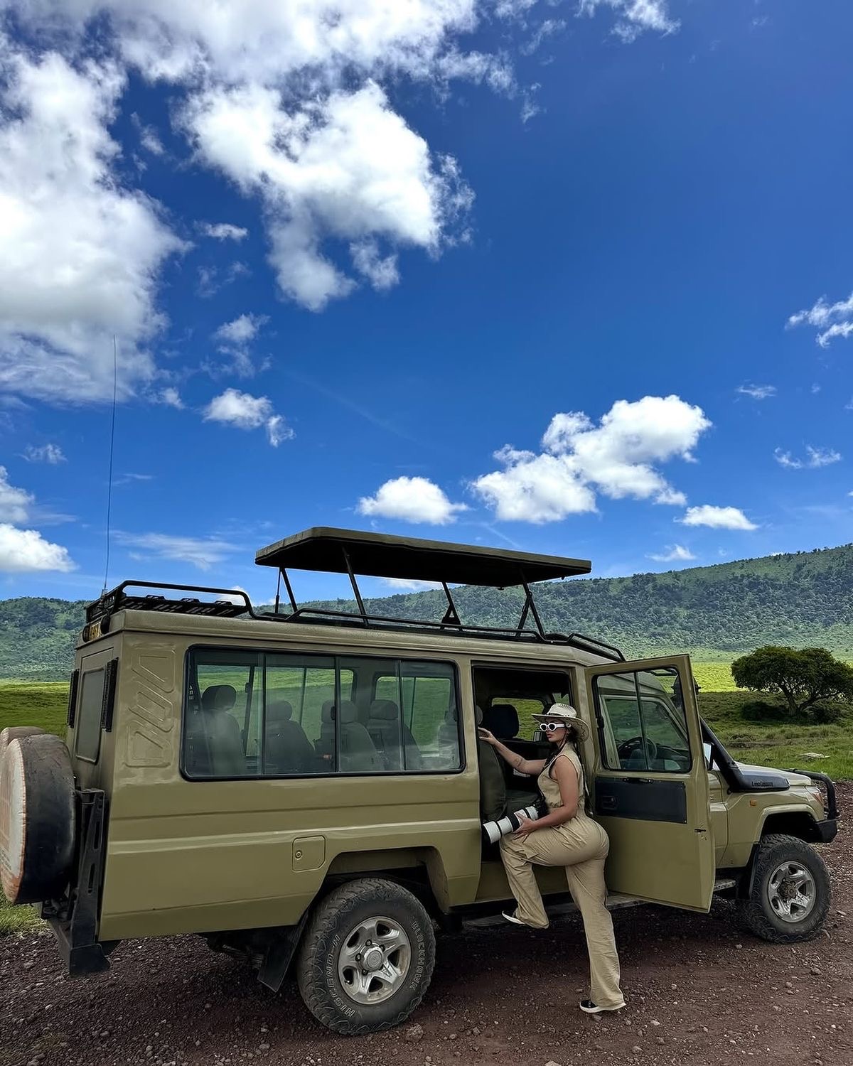 Traveller stepping into an open-roof safari Land Cruiser on the green Ngorongoro Crater rim under a bright blue sky.