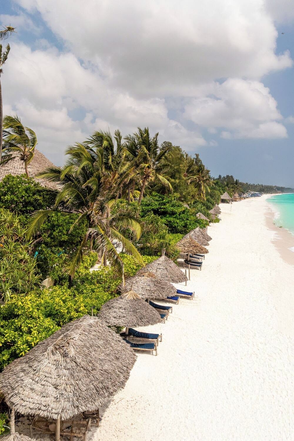 Aerial sweep of Nungwi's white-sand beach lined with thatched parasols and lush palm forest on Zanzibar's north coast.