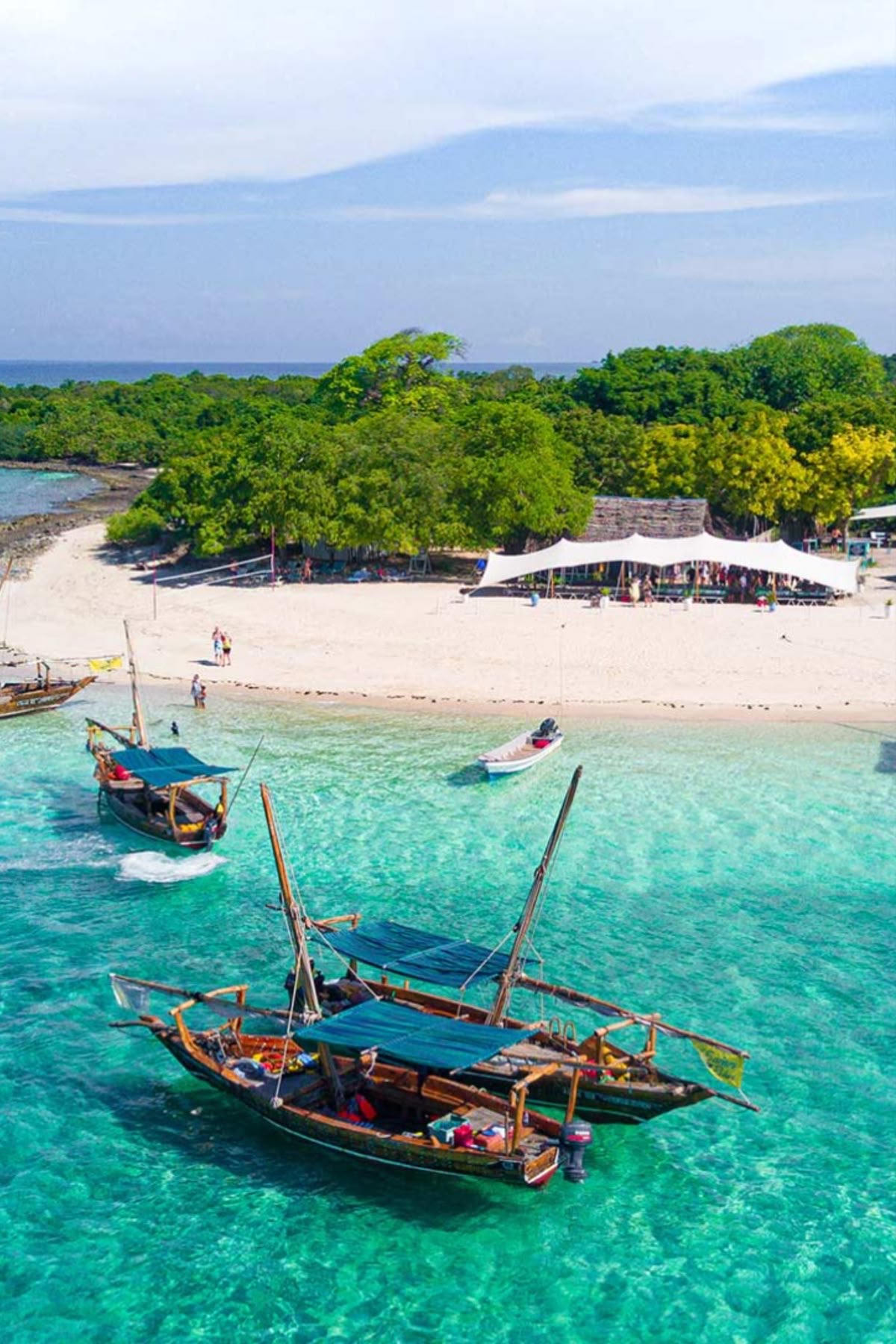 Aerial view of traditional dhows anchored beside a deserted sandbank with snorkelers wading in clear turquoise water.