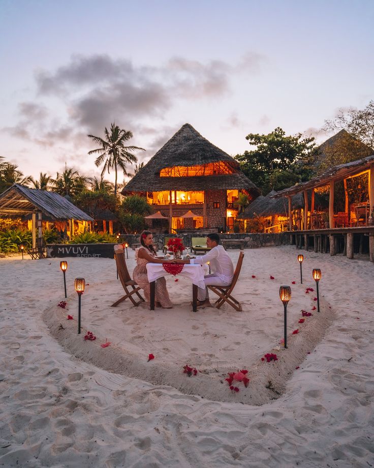 Romantic candlelit beach dinner for two on white sand in front of a thatched Zanzibar boutique resort at sunset.